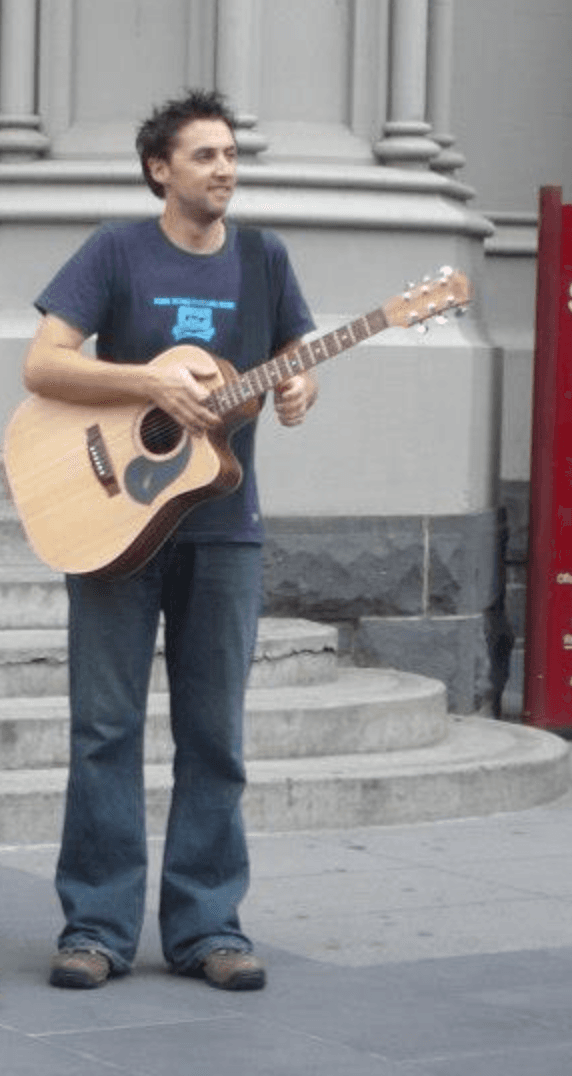 Ben stands on a city street playing an acoustic guitar and busking in front of a stone building.