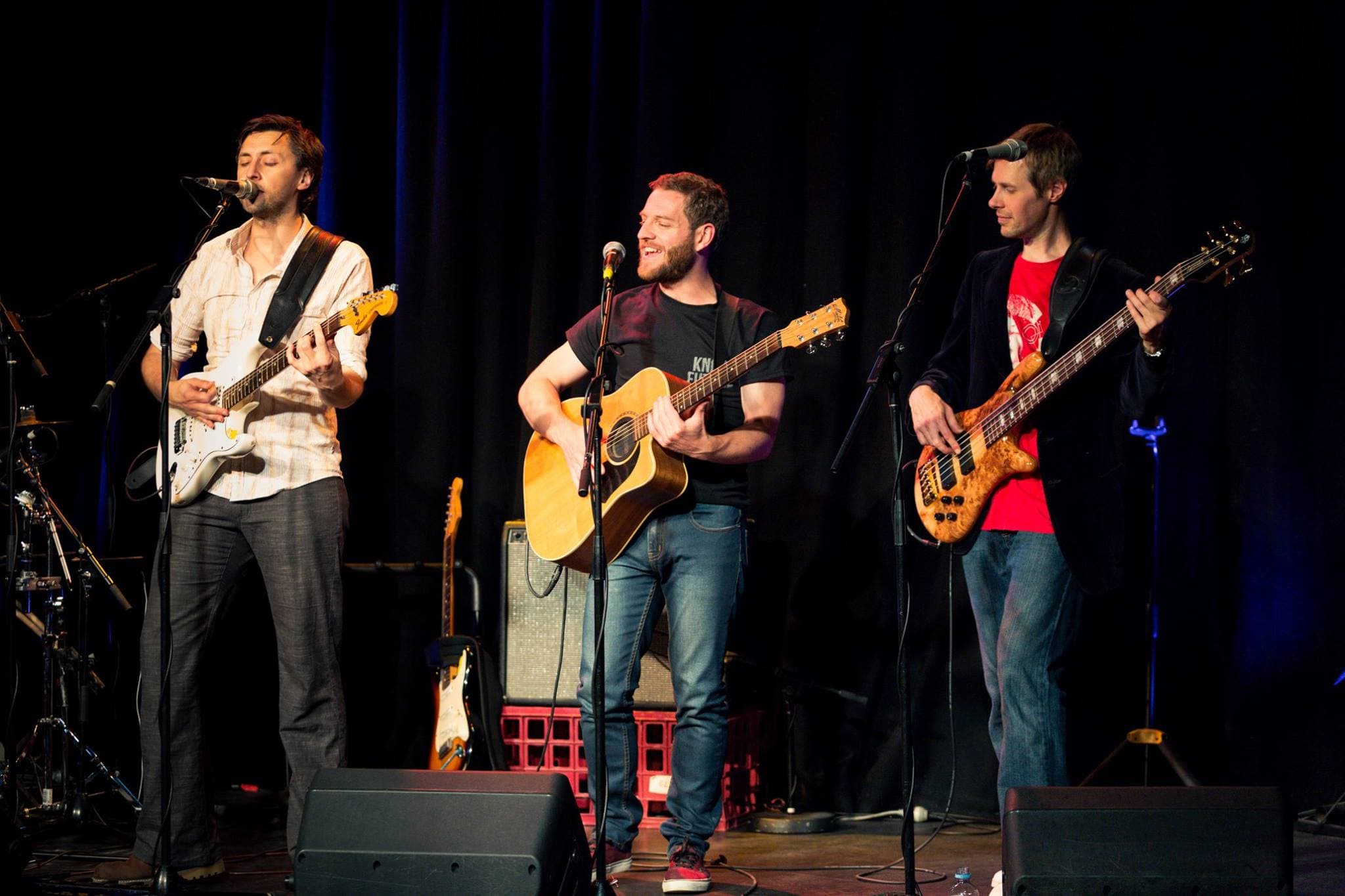 Ben sings into the mic while playing a white electric guitar, flanked by Shane on acoustic guitar and a bandmate on bass during a live stage performance.