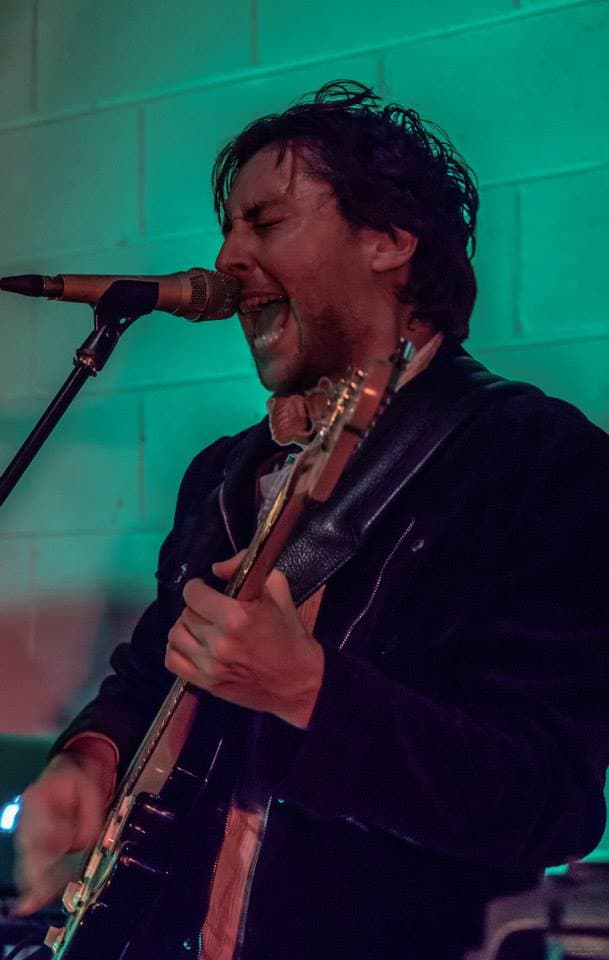 Ben sings hard into the mic with eyes shut while playing guitar under green stage light at a live show.