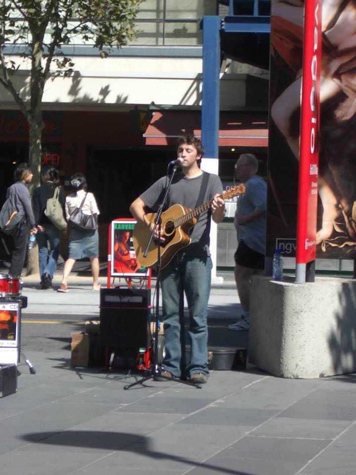 Ben busks in a wide city plaza, singing and playing acoustic guitar as pedestrians pass behind him.