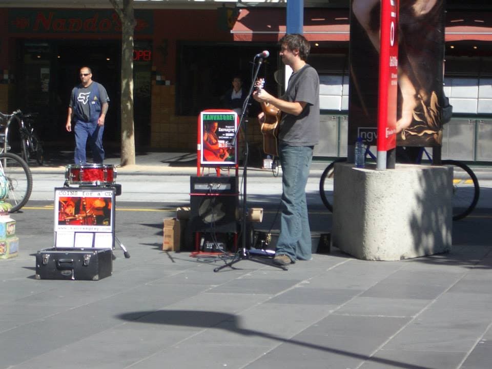 Wide shot of Ben busking with acoustic guitar in a sunlit pedestrian street while a man watches nearby.