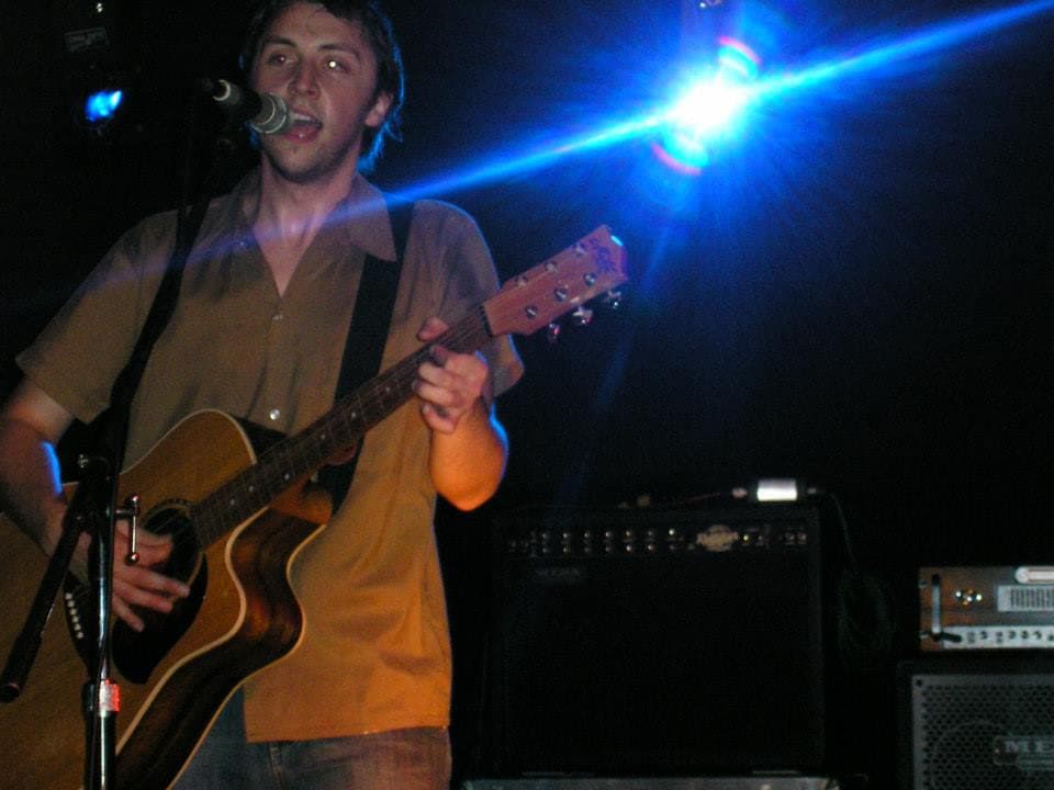 Ben sings into the mic and plays acoustic guitar onstage as a blue stage light flares behind him.