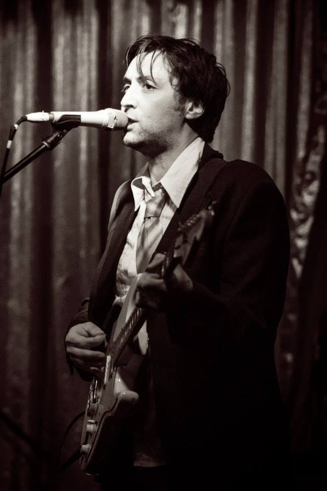 Ben sings into a microphone while playing electric guitar onstage in a sepia-toned live shot.