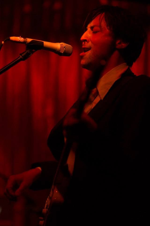 Ben sings and plays guitar under crimson stage lighting in front of a red curtain.