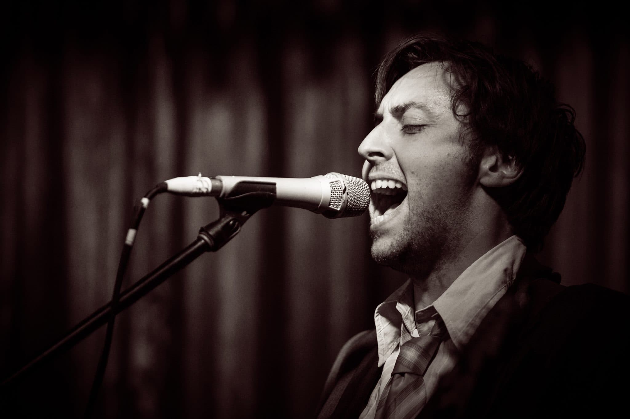Tight black-and-white close-up of Ben belting into the microphone with mouth wide open.