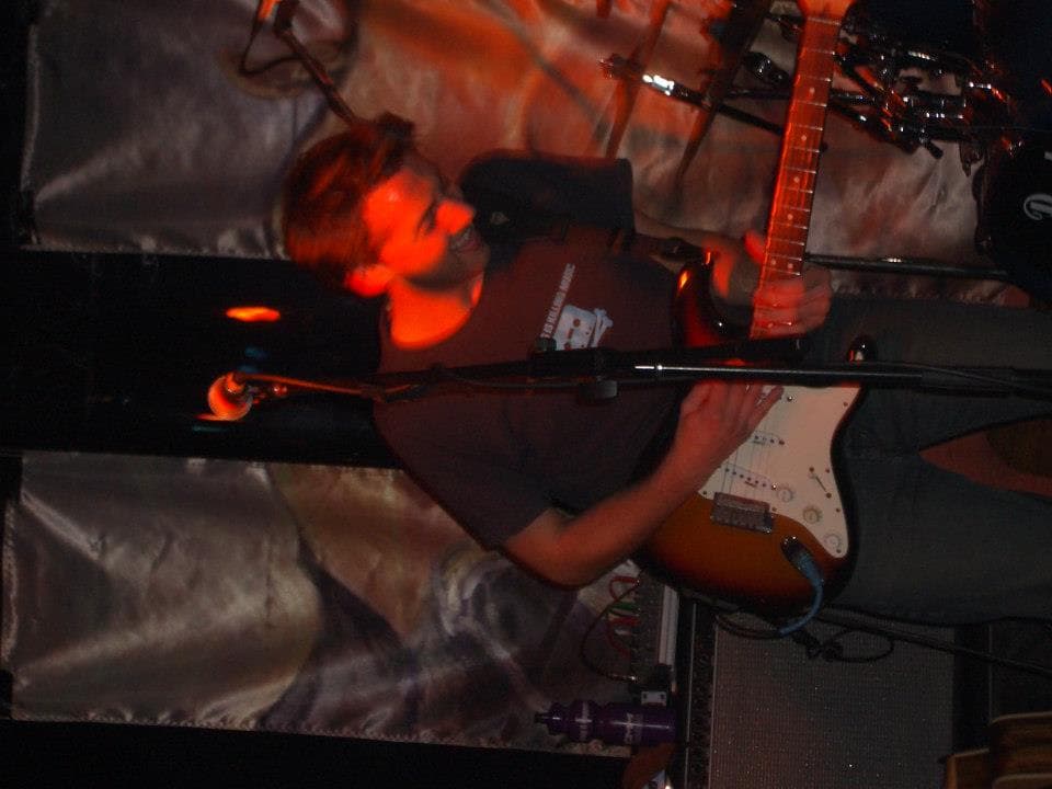 Ben sings into a microphone while playing a sunburst electric guitar under red stage lighting at a live show.
