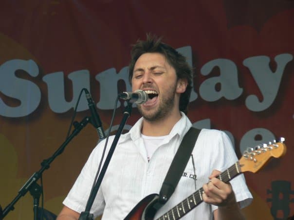 Ben sings open-mouthed into a microphone with his electric guitar in front of a colourful festival backdrop.