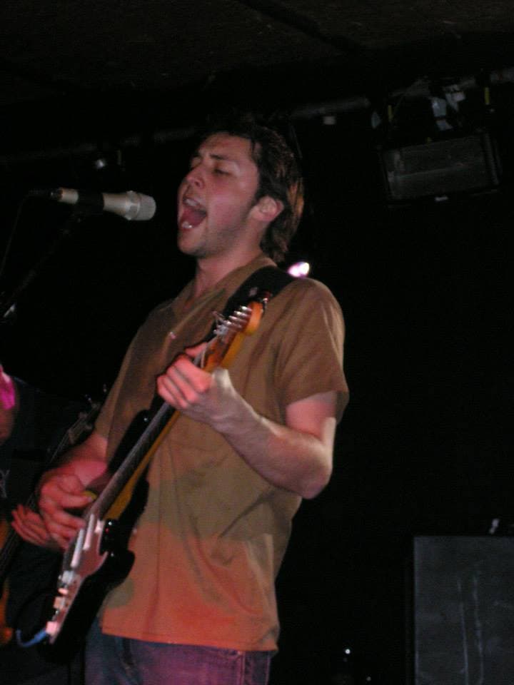 Ben sings passionately while playing electric guitar on a dark club stage.