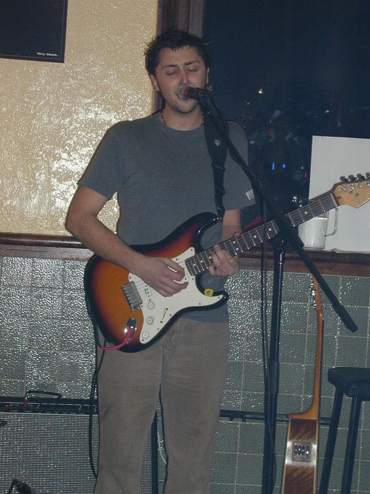 Ben sings into a mic with eyes closed while playing a sunburst electric guitar at an intimate pub gig.