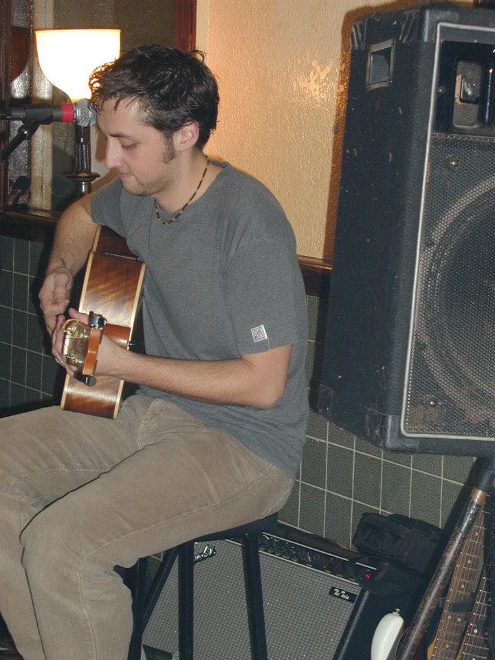 Ben looks down at his acoustic guitar as he plays, seated beside a stage monitor with a lamp glowing behind him.