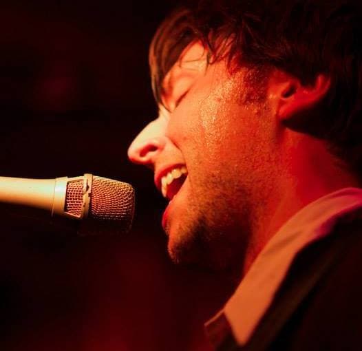Tight close-up of Ben singing into a mic with eyes shut, his face lit by warm red stage light.