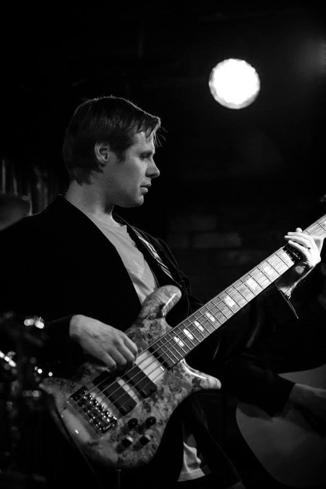 Black and white shot of Liam in profile playing his five-string bass beneath a stage light.