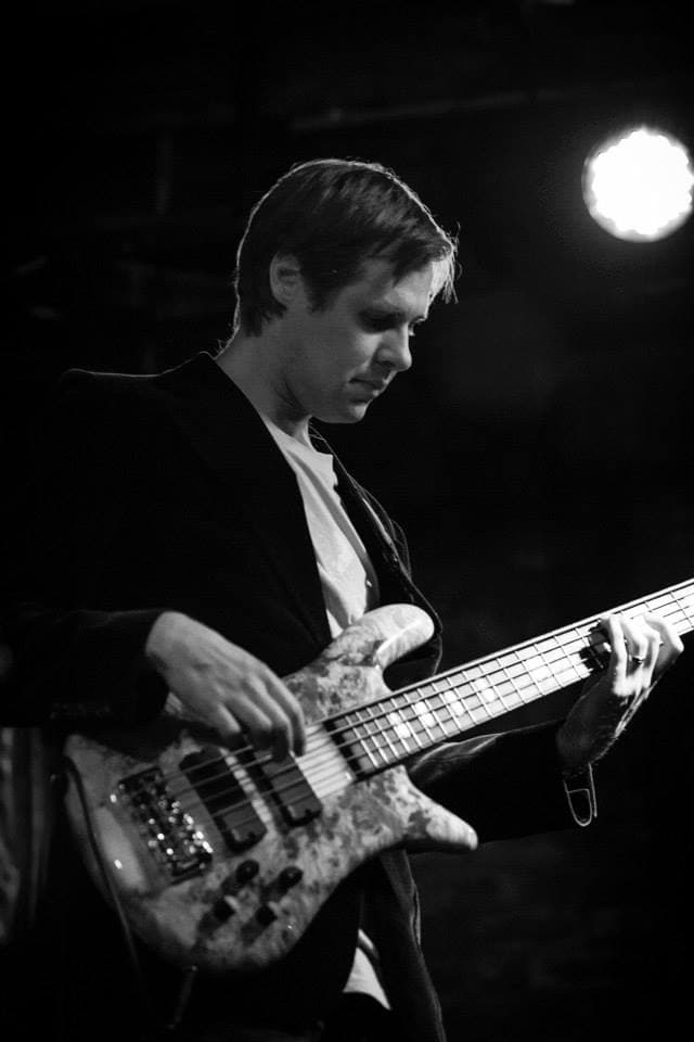 Black and white image of Liam looking down as he plays his bass under a stage spotlight.