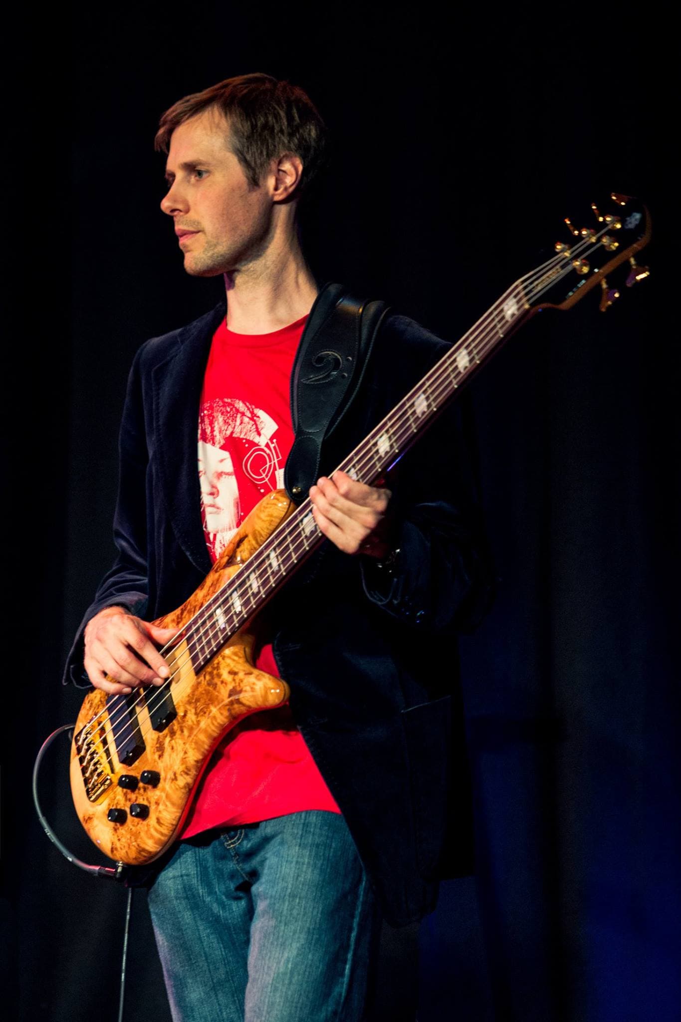 Liam stands on a darkened stage in a red shirt and blazer, holding his natural-wood bass and looking off to the side.