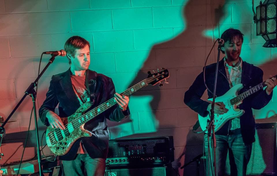 Liam on bass and Ben on guitar perform side by side under teal lighting against a brick wall.