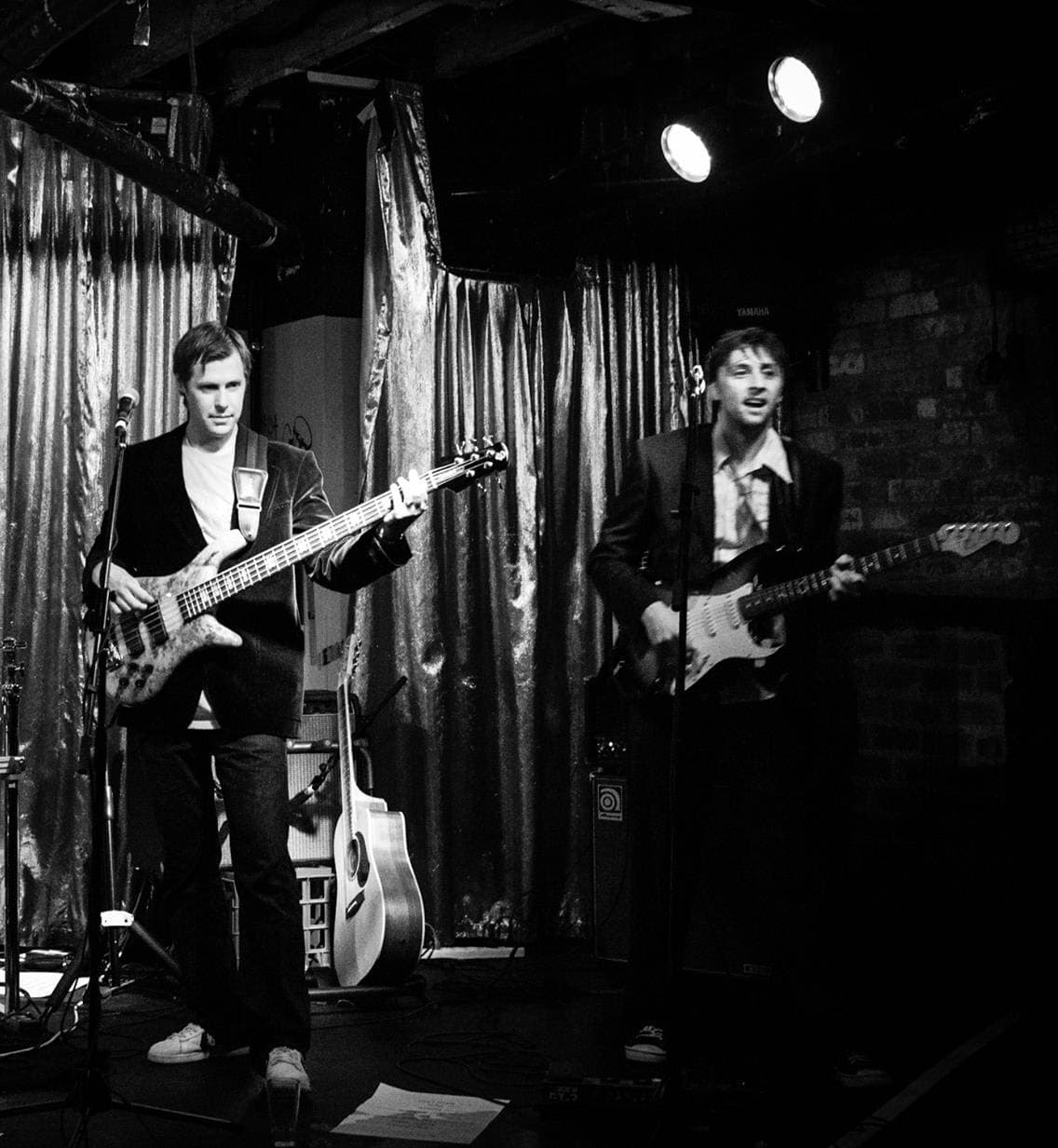 Black and white shot of Liam on bass and Ben singing with a guitar on a small stage with draped curtains.