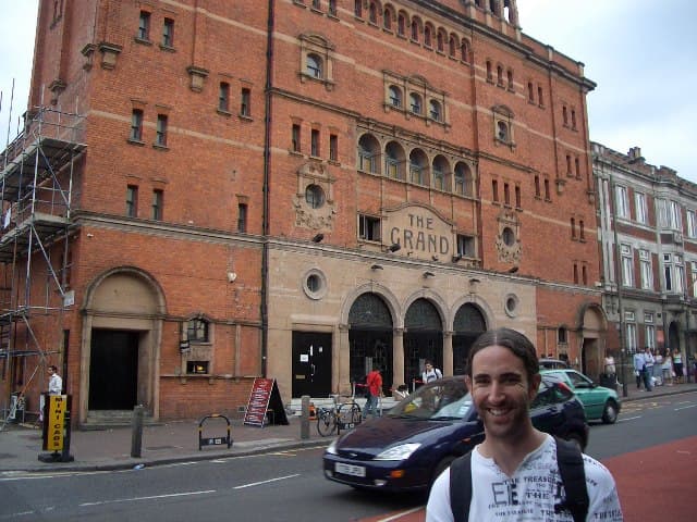 A long-haired band member smiles on the street in front of a red-brick building marked The Grand.