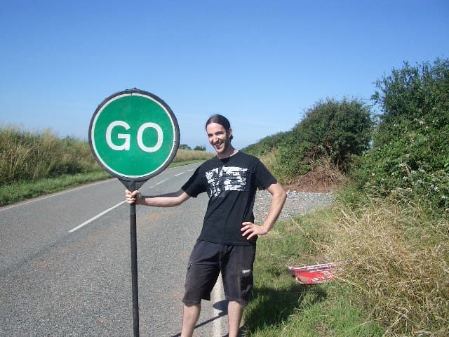 A long-haired band member grins on a country roadside next to a green road sign reading GO.