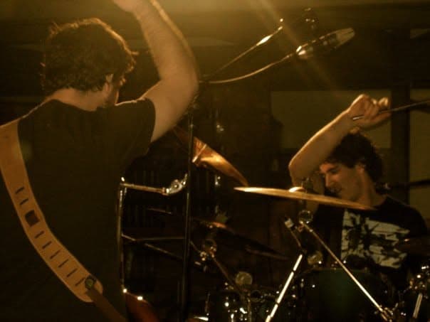 A guitarist with his arm raised plays beside Mark on drums under warm stage lighting at a live show.