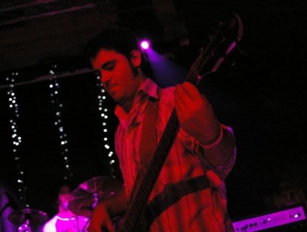 A bassist plays his bass head-down under deep pink and purple stage lighting at a live show.
