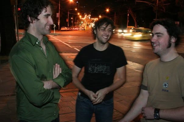 Three band members laugh together on a city sidewalk at night with traffic streaking past behind them.