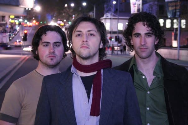 A three-member band portrait at night, the centre figure in a suit and scarf flanked by two bandmates against blurred city lights.