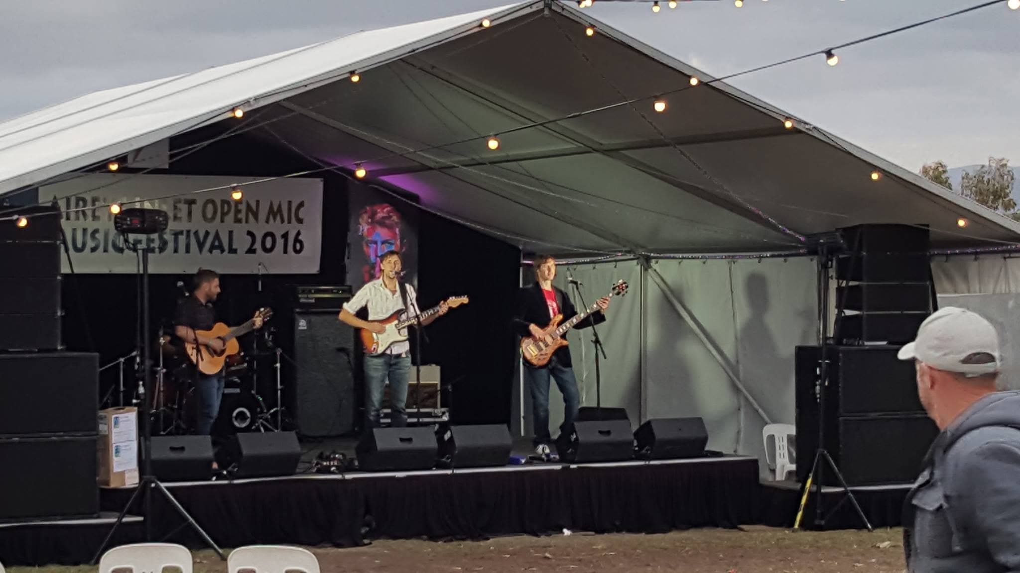 Kanvas Grey playing a marquee stage at the 2016 open-mic music festival, with an acoustic guitarist, a lead guitarist at the mic and a bassist under string lights.