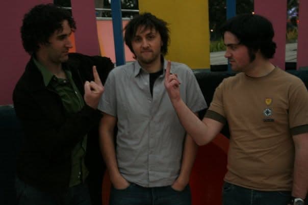 Three band members stand together against a colourful interior wall, the central one and another playfully pointing fingers in the air.
