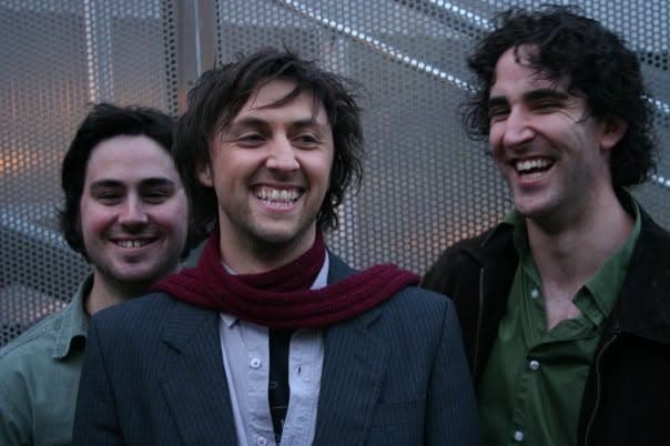Three band members laughing together outdoors in front of a metallic mesh wall, the central one wearing a dark red scarf.