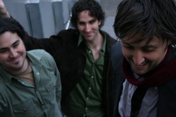 Three band members photographed from below outdoors, two looking down and grinning while a third smiles in the background.