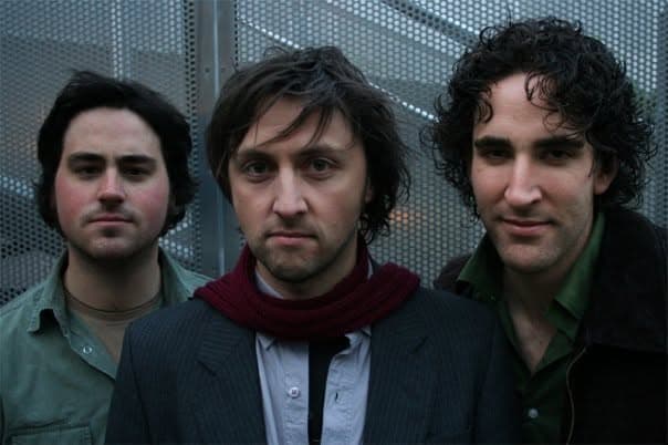 Three band members pose with serious expressions against a metallic mesh wall, the central one wearing a dark red scarf and grey jacket.