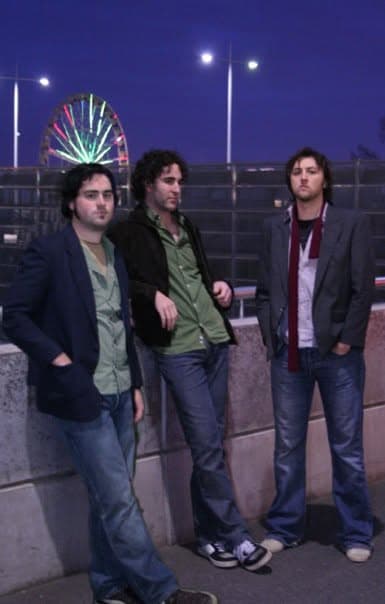 Three band members stand on a rooftop or boardwalk at dusk with a brightly lit Ferris wheel glowing behind them.