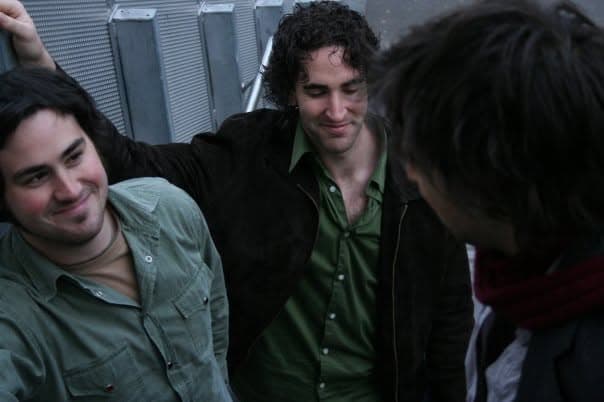 Three band members sit close together against a metal wall, two smiling toward a third whose back is to the camera.