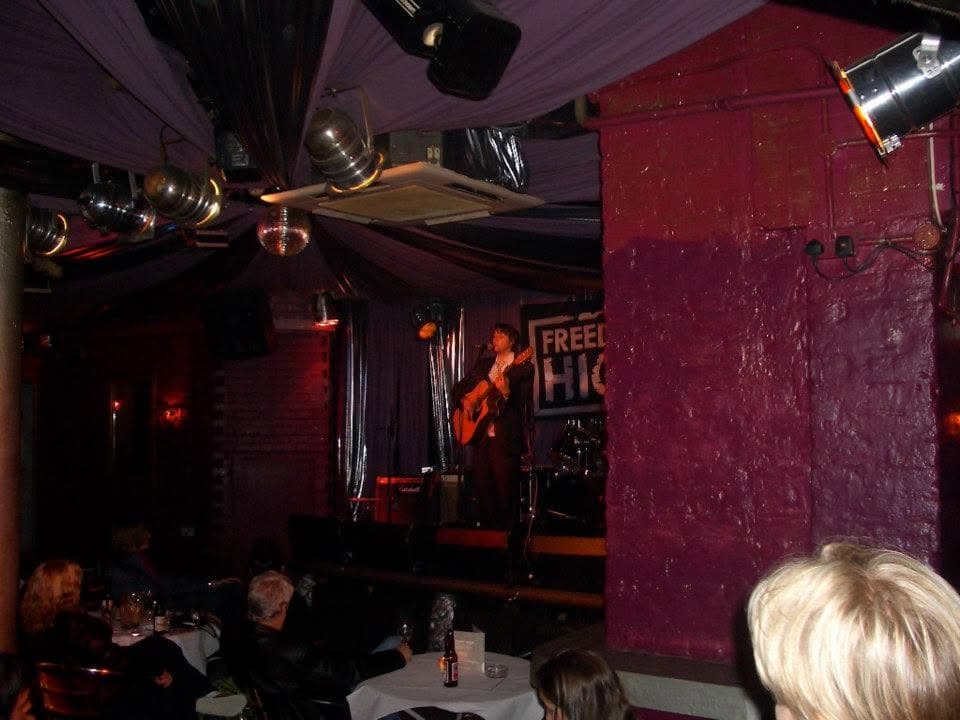 Ben Thomas sings and plays acoustic guitar alone on a small purple-draped club stage in front of a 'Freedom High' banner, with seated patrons at candlelit tables in the foreground.