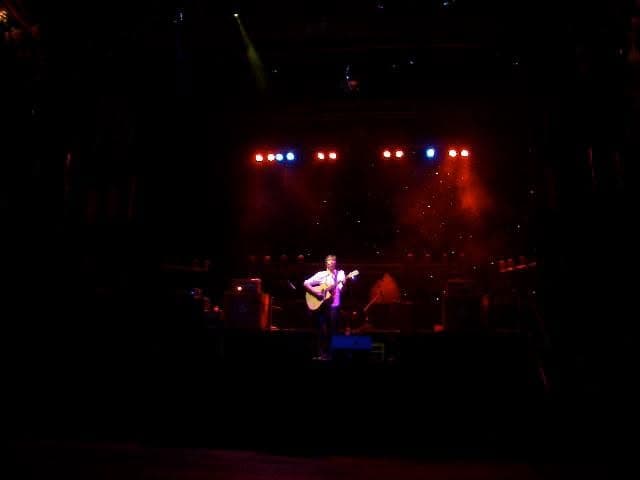 A lone musician with an acoustic guitar stands centre stage on a large dark venue stage lit by red and blue spotlights.