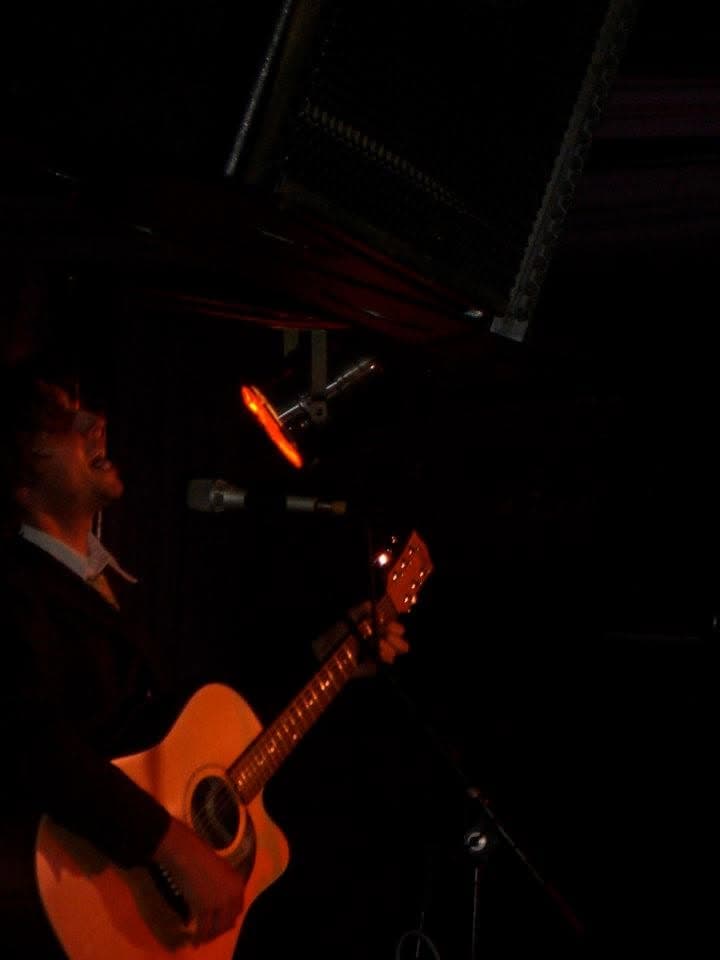 Ben Thomas sings into a microphone while playing an acoustic guitar in a dark, dimly red-lit venue.
