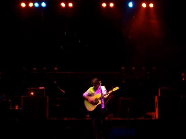 A solo guitarist performs centre stage under a row of red and blue stage lights in a large darkened venue.