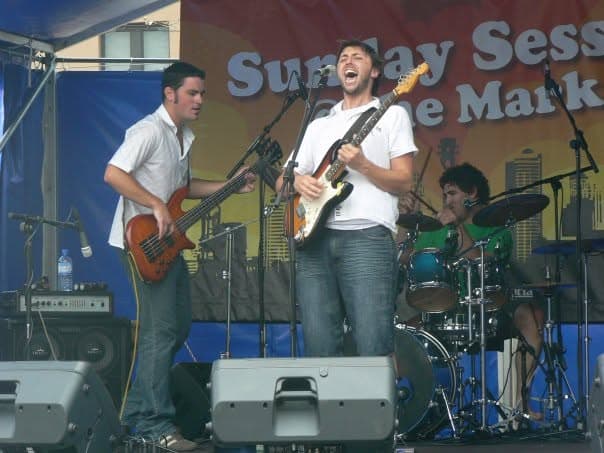 Ben Thomas sings wide-mouthed into the mic while playing guitar at an outdoor festival, with a bassist beside him and the drummer behind, under the 'Sunday Sessions @ the Market' banner.