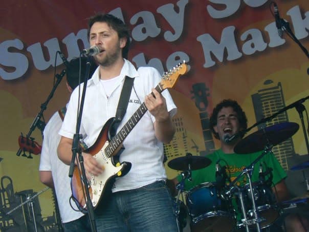 Ben sings into a microphone while playing a sunburst electric guitar onstage as a bandmate drums in the background at an outdoor Sunday Sessions market gig.