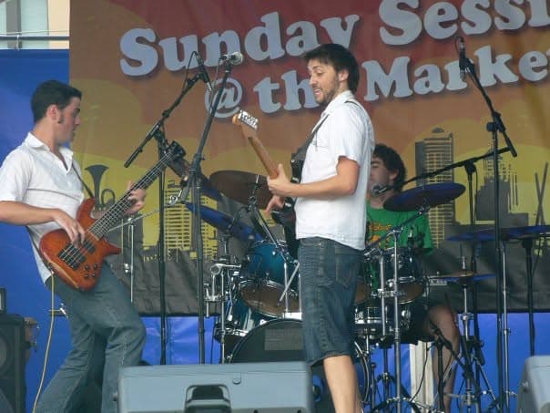 The band performs onstage with Liam on bass at left, Ben center on guitar, and the drummer behind at the outdoor Sunday Sessions market stage.