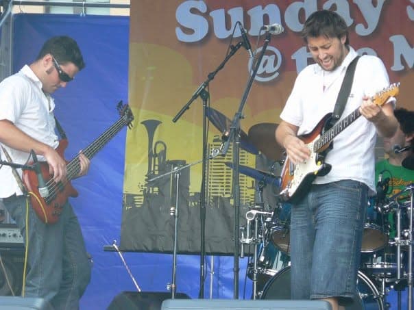 Liam plays bass in sunglasses on the left while Ben leans into a guitar solo on the right during an outdoor Sunday Sessions performance.
