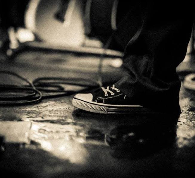 A close-up black-and-white shot of a worn Converse sneaker on a scuffed stage floor beside a coiled cable.
