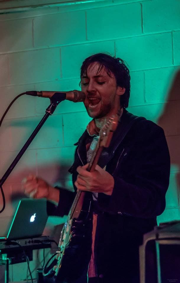 Ben sings eyes-closed into the mic with an electric guitar against a teal-lit brick wall, a glowing Apple laptop on the floor beside him.