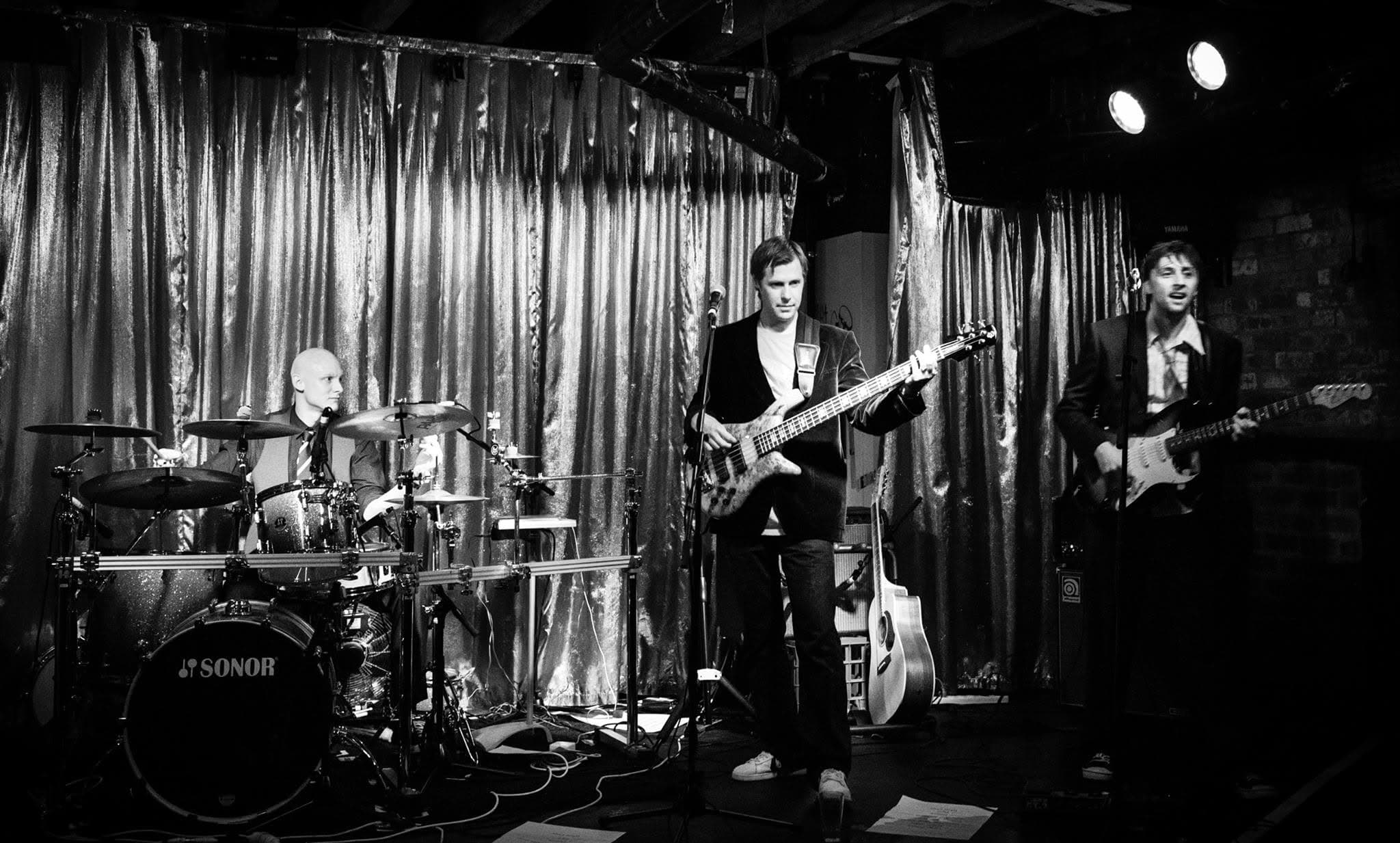 Black and white: drummer in a suit and tie at a Sonor kit, bassist centre in a jacket, singer with a Stratocaster on the right, all in front of a shimmering curtain under two spotlights.