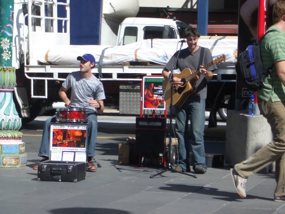 Busking on a city footpath: a drummer in a cap on a snare and cymbal, the singer with an acoustic guitar, and a hand-lettered 'Coins for a CD' sign with the band's web address propped on an open case.