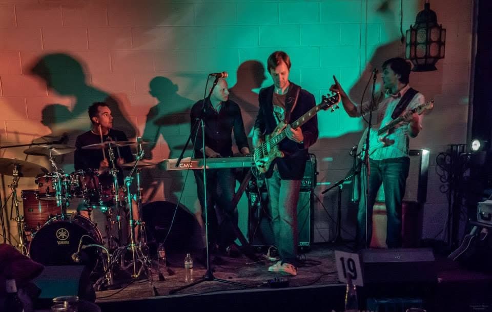 Four-piece against a cinderblock wall washed in teal and orange light: drummer at a Yamaha kit, keys, bassist centre in a blazer, and a guitarist pointing skyward, their shadows thrown up the wall.