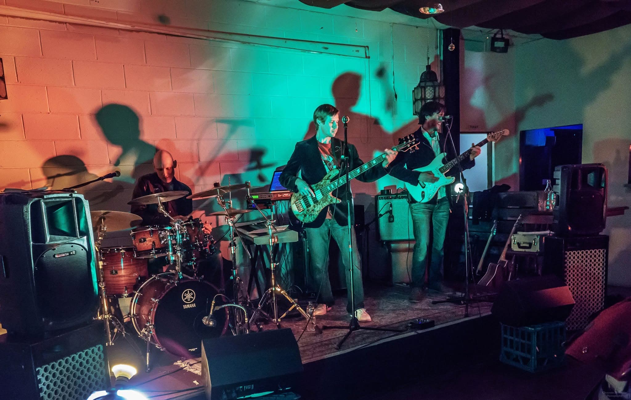 Wide shot of the colour-washed room: drummer, keys and a laptop, bassist centre, guitarist with a white Stratocaster on the right, PA cabinets and a milk crate in the foreground, shadows across the cinderblock wall.