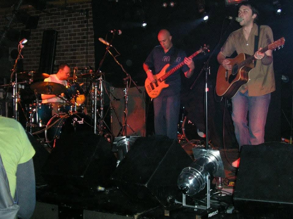 On a brick-walled stage: drummer in a white T-shirt, bassist in a dark shirt, and the singer at the mic with an acoustic guitar, wedge monitors along the front of the stage.