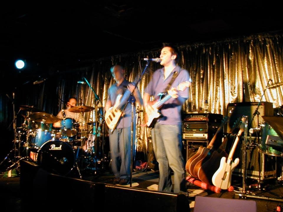 Singer at the mic with a Stratocaster under stage light, bassist and drummer behind, spare guitars on stands at the side, a silver tinsel curtain across the back of the stage.