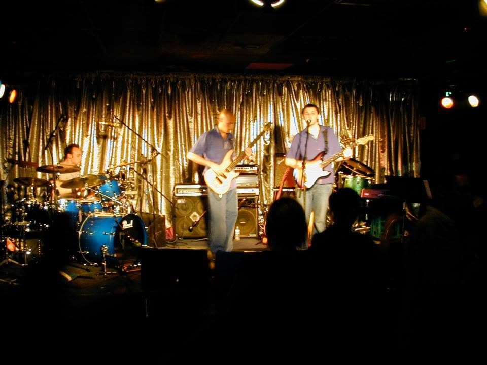 Three-piece on a club stage in front of a silver tinsel curtain: drummer at a blue Pearl kit on the left, bassist centre, singer with a Stratocaster at the mic, silhouettes of the crowd in the foreground.